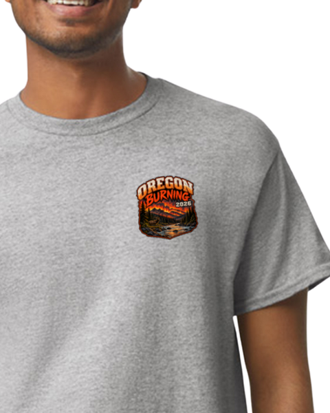 A man wearing a heather gray crewneck t-shirt with a small chest graphic that reads 'OREGON BURNING 2026' above an orange and yellow landscape illustration.