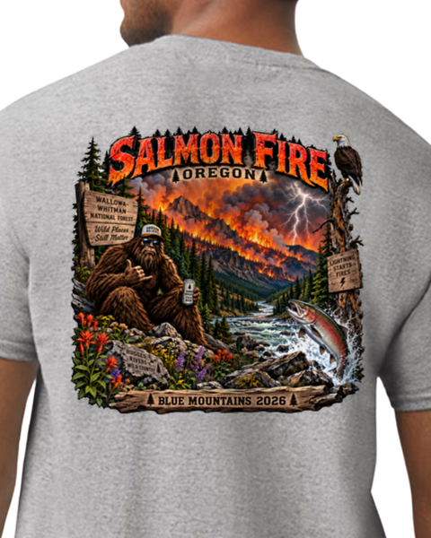 A man wearing a heather gray t-shirt, viewed from the back. The shirt features a large, detailed graphic print of a Bigfoot scene in a volcanic Oregon landscape with the text 'Salmon Fire' and 'Blue Mountains 2026'.