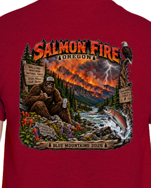 A close-up of a red t-shirt showing a large graphic print on the back. The graphic features a Bigfoot scene in a forest with a river, a jumping salmon, lightning, and a volcano erupting under the text 'SALMON FIRE OREGON' and 'BLUE MOUNTAINS 2026'.