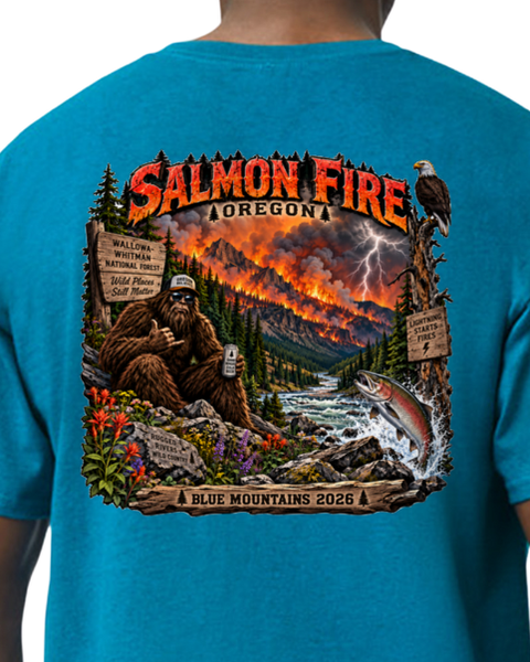 A close-up of the back of a person wearing a bright blue t-shirt. The shirt features a large, detailed graphic print of a Bigfoot scene in a forest fire landscape with a salmon jumping in a river, and the text 'SALMON FIRE OREGON' and 'BLUE MOUNTAINS 2026'.