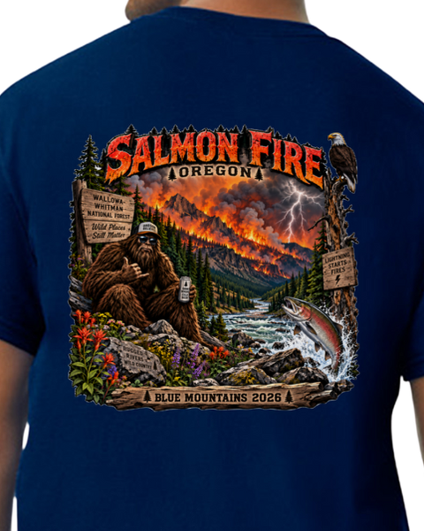 A navy blue t-shirt featuring a large, detailed back graphic of a Bigfoot scene in Salmon Fire, Oregon, with a Bigfoot sitting by a river, a jumping salmon, lightning, and a mountain landscape. The text 'SALMON FIRE OREGON' is prominent at the top, and 'BLUE MOUNTAINS 2026' is at the bottom.