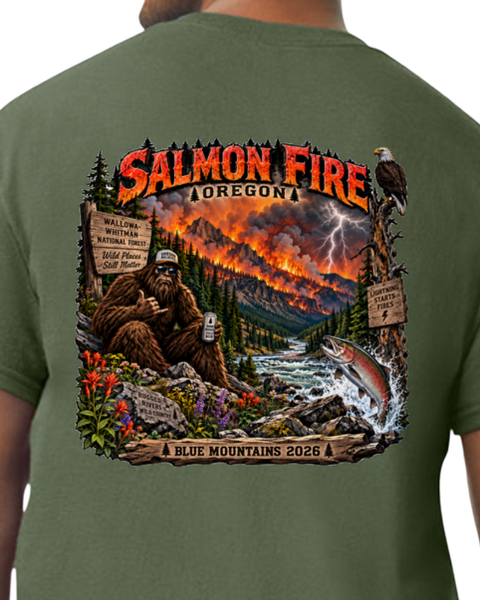 A close-up of a person wearing an olive green t-shirt, showing the back of the shirt with a large, detailed graphic print. The graphic features a Bigfoot scene in a forest with a river, a jumping salmon, a lightning storm, and the text 'SALMON FIRE OREGON' and 'BLUE MOUNTAINS 2026'.