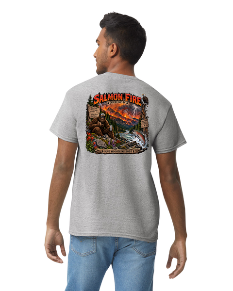 A man wearing a heather gray t-shirt with a large, colorful graphic print on the back depicting a Bigfoot mountain wildfire scene with the text 'SALMON FIRE' and 'BLUE MOUNTAINS', paired with blue jeans, shown against a plain white background.