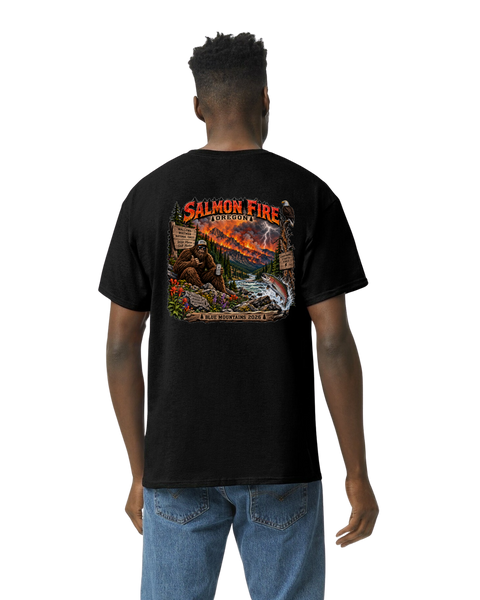A man viewed from the back wearing a black t-shirt with a large graphic print on the back. The graphic features the text 'SALMON FIRE' in red and orange, above an illustration of a river, mountains, wildfire and a bigfoot.